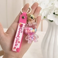 Nahaufnahme von rosa Bär-Schlüsselanhänger mit Good Luck Band und goldener Glocke - Rosaroter Bär mit Glocke