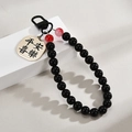 Black beaded keychain with gold metal tag engraved Ping An Xi Le on white podium - Black Peace And Joy