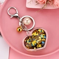 Détail de porte-clés cœur acrylique avec paillettes, clochette et perle sur fond floral rose - Doré