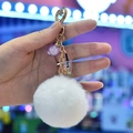 Detail of fluffy white fur pompom keychain with purple bead and heart-shaped gold clasp - White