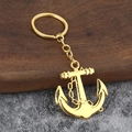 Polished golden metal anchor keychain with circular ring on rustic wooden and stone background - Gold