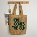 Bolso tote de paja con bordado verde Here Comes The Sun colgado de un gancho - Marrón