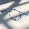 Macro shot of blue evil eye woven bracelet with gold bead on textured white surface - Gold-Sky Blue Rope