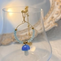 Detail eines blauen Pilz-Ohrrings mit goldenem Ring und hellblauen Perlen an einem Glas - Blau