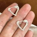 Detalle de aretes de corazón plateados sostenidos a mano sobre superficie de felpa - Plateado