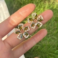 Macro detail of olive green zircon flower earrings held on fingers with outdoor background - Olive Green