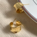 Dettaglio macro di orecchini larghi a C dorati con trama ondulata su tessuto beige - Oro