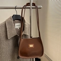 Bolso de hombro en cuero marrón con logo dorado colgado frente a un abrigo de cuadros - Marrón