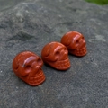 Three hand-carved natural red jasper skull figurines displayed on a grey rock surface - Red Jasper