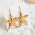 Dettaglio di orecchini a stella marina dorati su cartoncino bianco su sabbia di spiaggia - Stile Stella Marina d'Oro (29422-2)