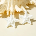 Detailed macro view of white starfish resin earrings with realistic texture on a beach setting - White