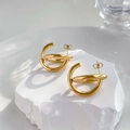 Detail van gouden C-vormige crossover-oorbellen op een wit gestructureerd stenen oppervlak - Kruis-C-stijl / Goud