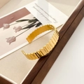 Dettaglio di un bracciale in acciaio inossidabile dorato con trama a onde su sfondo vintage - Polsino Aperto a Ingranaggio Largo (Oro)