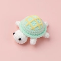 Top-down view of a handmade crochet turtle plush toy with green shell on pink background - Blue Biscuit (Turtle Doll)