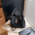 Schwarze Eimer-Tasche mit Litschistruktur, Goldschloss und Gurt vor einem Trenchcoat - Schwarz