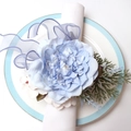 Detail eines hellblauen Sonnenblumen-Serviettenrings mit Perle und Band auf dem Teller - Rose / Blau