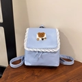 Dettaglio dello zaino azzurro con bordo in pizzo bianco e toppa con orsetto di peluche - Blu
