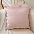 Pink velvet pillow cover with pom-pom trim displayed on a soft beige fabric sofa - Pink