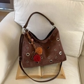 Detail der braunen Wildleder-Hobo-Tasche mit Metallösen und Pompons auf einem Sofa - Kaffee Mit Pompon-Schlüsselanhänger