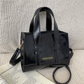 Glossy black square shoulder bag with gold lettering on a striped fabric background - Black