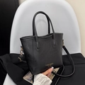 Bolso tipo cubo negro con textura de cuero y letras doradas sobre silla blanca - Negro
