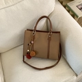 Brown woven tote bag with adjustable shoulder strap and colorful pom-pom charm on sofa - With Charm / Brown