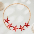Macro detail of gold body chain featuring vibrant red starfish charms on sandy beach decor - Gold+Red
