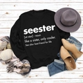 Flat lay van een zwart sweatshirt met witte Seester-opdruk op een houten ondergrond - S / Zwart (Wit Lettertype)