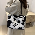 Furry cow print tote bag with black leather straps shown on shoulder in a cozy room - Black