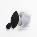 Detalle de dos esponjas de maquillaje negras con corte de oliva y bolsa esmerilada - Corta Una Aceituna Negra En Rodajas Y Añade Una Mini Gota De Agua (en Una Bolsa Escarchada).