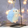 Close-up of blue hedgehog plush keychain hanging on a metal stand with bokeh background - Height about 11 / Light Blue