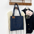 Dark blue denim tote bag with leather patch and outer pocket hanging on a wooden rack - Style 1 (With Front Pocket)