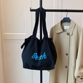Black canvas tote bag with blue Beach text and drawstring closure on clothes rack - Black
