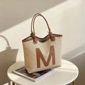 Minimalistische beige Web-Tote-Bag mit braunem Leder-M-Logo auf einem Beistelltisch - Khaki