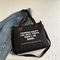 Black canvas tote bag with white lettering and adjustable shoulder strap shown on denim jacket - Black