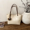 Detalle de bolso tote de cuero crema con asas marrones y colgante en banco de madera - Con Colgante / Blanco