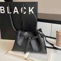 Vista detallada del bolso trapezoidal negro mate con lazo decorativo en un entorno de ciudad - Negro