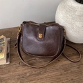 Bolso bandolera vintage marrón oscuro con hebilla dorada sobre mesa de madera - Marrón