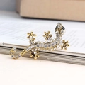 Broche gecko dorée ornée de strass blancs et yeux en pierre noire sur un livre - Petit Blanc