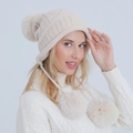 Close-up of cream white knit beanie with soft plush pom poms and ear flaps for winter comfort - Beige