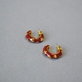 Detail von roten Emaille-C-Ohrringen mit goldenen Herzmustern auf grauem Stoff - Rot