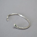 Macro detail of silver snake bracelet showing scales and green gemstone eyes on snake head - Silver