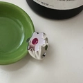 Anillo de flor minimalista en plata S925 con piedra roja sobre una mesa blanca - Abierto Ajustable / Pequeña flor de plata con circonita roja (JZA081) / Anillo Ajustable. La Superficie Del Anillo Presenta Un Sencillo Diseño De Flor De Cinco Pétalos Hecha De Metal Plateado Con Una Gema De Color Púrpura Rojizo (visiblemente Un Circón Rojo) Engastada En El Centro De La Flor.
