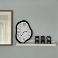Melting surrealist wall clock with black frame and roman numerals on a white shelf - Black and White
