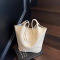 Minimalistische beige Zylindertasche mit Lychee-Textur und dünnen Griffen auf dunkler Treppe - Cremeweiß