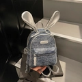 Blauwe denim mini rugzak met zilveren konijnenoren en modelabel, op een boek gezet - Denim Blauw