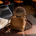Braune Beuteltasche mit geflochtenem Griff und Goldlogo in einer gemütlichen Café-Kulisse - Braun