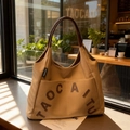 Khaki canvas tote bag with brown leather handles and letter patch in a cafe setting - Caramel color