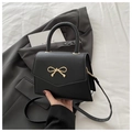 Detalle de bolso trapezoidal negro con hebilla de lazo dorado sobre fondo de blazer gris - Negro