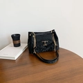 Black pleated leather shoulder bag with gold T-buckle on a minimalist wooden table - Black